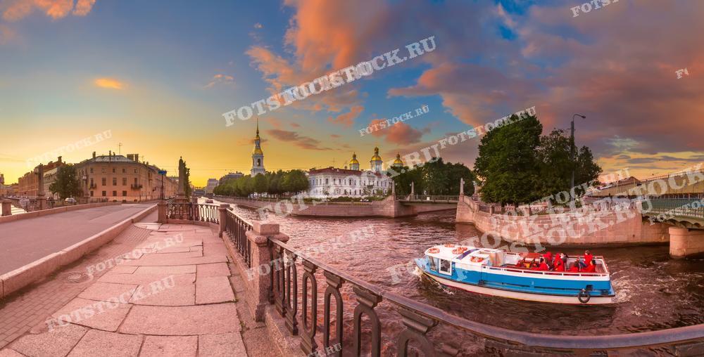 На переднем плане этой фотографии расположено кованое ограждение набережной в Петербурге, а в центре композиции — проплывающий экскурсионный катер с туристами. На заднем плане видны архитектурные достопримечательности города. Такое фото отлично подойдёт для публикаций о путешествиях, для промо-материалов туристических агентств или как красочный элемент украшения интерьера.